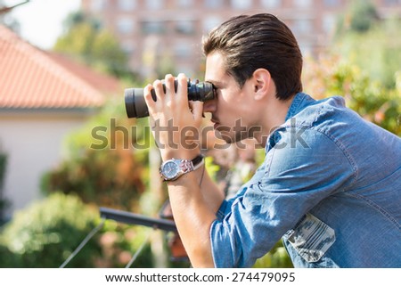 Young man looking through binocular searching outside from balcony