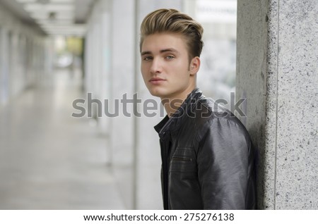 Attractive blond young man with leather jacket standing outside against pillar, looking down at camera
