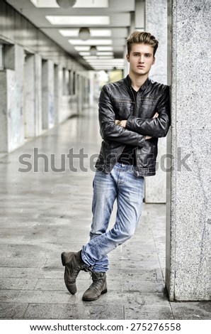 Attractive blond young man with leather jacket standing outside against pillar, looking at camera