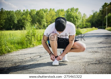 Handsome young man tying sports shoes before going running and jogging on road in the country, wearing white shirt and baseball cap