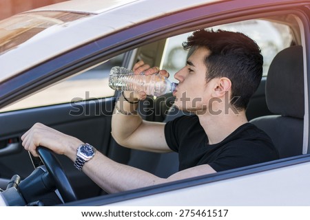 Handsome young man or teenager driving car and drinking water from plastic bottle