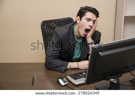 Tired bored young businessman sitting at his desk in front of his computer yawning, with his chin resting on his hand and eyes closed, in his office