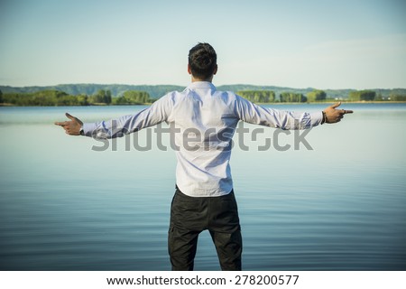 Young man in shirt, outdoor with arms spread open enjoying freedom in front of lake, seen from the back.