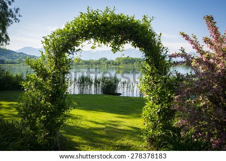 Woven plants forming an arch in beautiful garden by the lake, surrounded by flowers