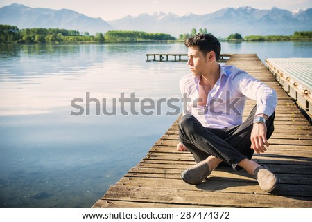 Handsome young man on a lake in a sunny, peaceful day, sitting on a wood pier, thinking or meditating