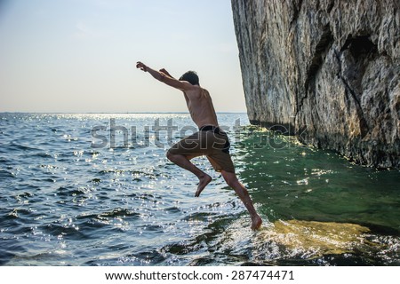 Attractive young shirtless athletic man jumping in water by sea or ocean shore, wearing shorts
