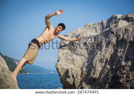 Handsome Shirtless Young Man Wearing Swim Shorts and Waving at Camera Leaning Across Gap Between Two Massive Boulders at Picturesque Ocean Coast
