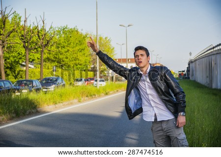 Handsome young man, a hitchhiker waiting for car on roadside in city, wearing black leather jacket