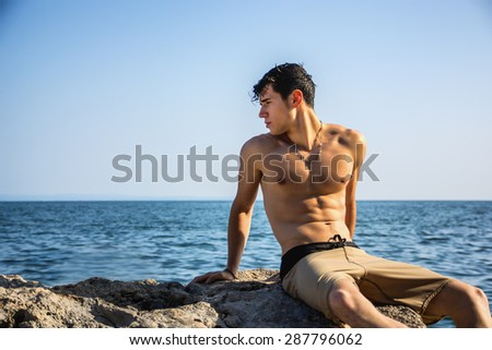 Attractive young shirtless athletic man crouching in water by sea or ocean shore, wearing shorts, looking away to a side
