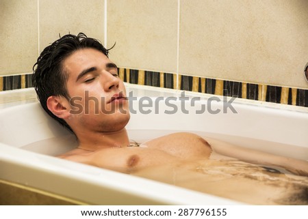 Handsome young man in bathtub at home having bath, washing body and hair with bathfoam and shampoo