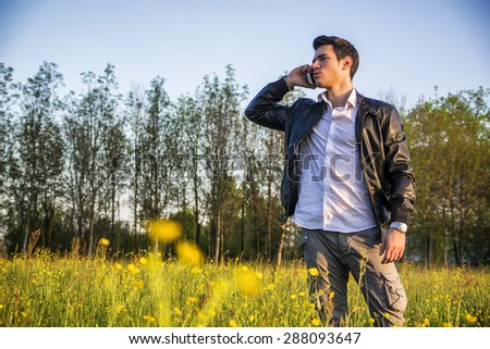 Handsome young man at countryside, talking on cell phone, standing in  field or grassland, wearing white shirt and jacket