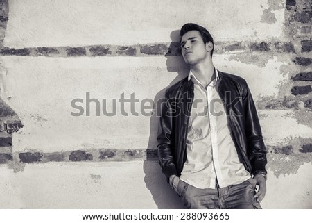 Handsome young man standing in white t-shirt and black leather jacket leaning against old wall behind him, looking away