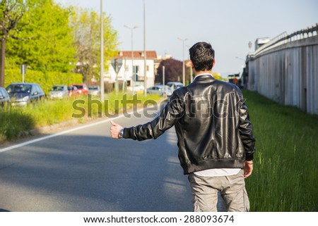 Back of young man, a hitchhiker waiting for car on roadside in city, wearing black leather jacket