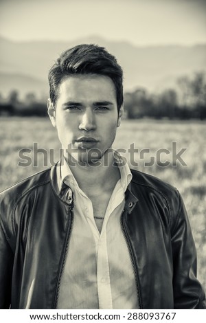 Handsome young man at countryside, in front of field or grassland, wearing white shirt and jacket, looking at camera