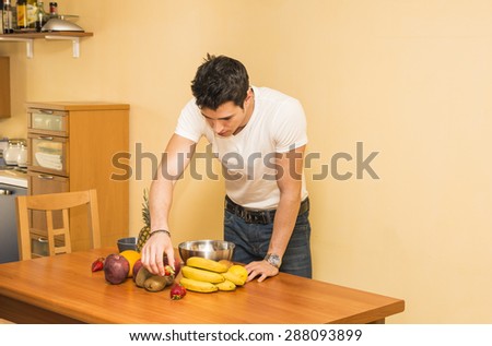Young man preparing a healthy fruit salad or smoothie, standing leaning on a kitchen counter loaded with an assortment of fresh tropical fruit, looking down