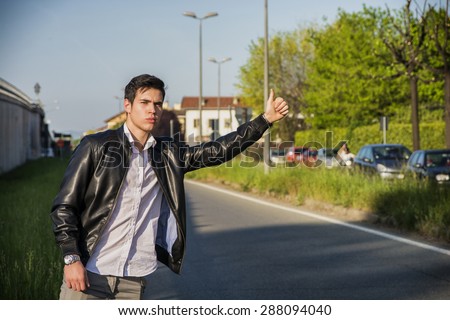 Handsome young man, a hitchhiker waiting for car on roadside in city, wearing black leather jacket