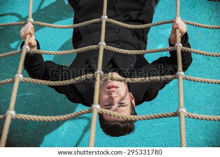 Full length of serious dark haired 20s man wearing black jacket and trousers lying upside-down on climbing rope, intensely looking at camera