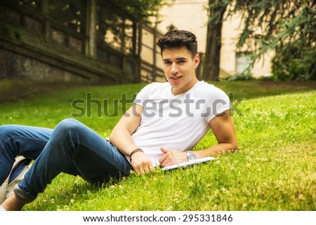 Young Male Student Studying his Lessons while Lying on Grass in City Park, Smiling at Camera