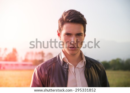 Handsome young man at countryside, in front of field or grassland, wearing white shirt and jacket, looking at camera