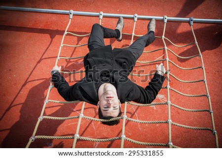 Full length of serious dark haired 20s man wearing black jacket and trousers lying upside-down on climbing rope, intensely looking at camera