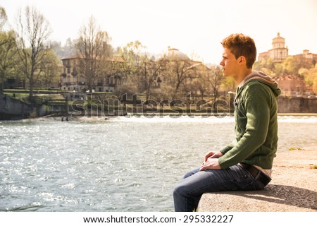 Three-quarter length of contemplative light brown haired teenage boy wearing green hooded-shirt and denim jeans sitting on wall beside picturesque river in Turin, Italy
