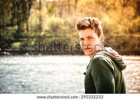 Head and shoulders length of contemplative light brown haired teenage boy wearing green hooded-shirt beside picturesque river or lake, looking at camera