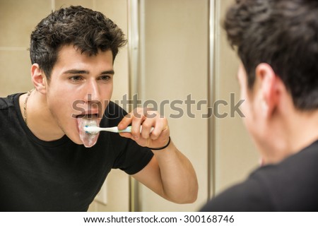 Headshot of attractive young man brushing teeth and cleaning tongue with toothbrush, looking at himself in mirror