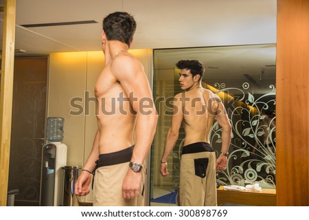 Young Shirtless Handsome Muscular Man Admiring His Muscles in Gym Mirror or Checking Progress and Training Results, as seen from Behind
