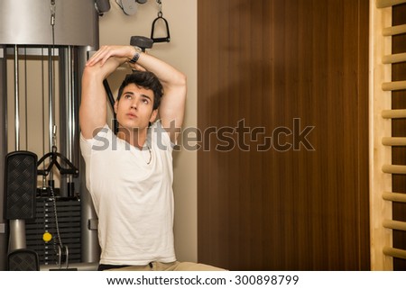 Attractive young man stretching arms in gym, touching foot with hands, looking in camera