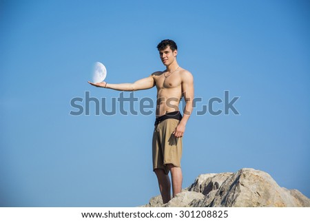 Muscular young man shirtless standing on rock against the sky with moon next to him, seen from below perspective