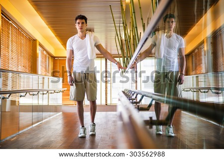 Low Angle View Full Length of Young Man with Towel Over Shoulder Standing in Bright Modern Hallway Holding on to Hand Rail