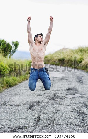 Shirtless sexy muscular young man jumping for joy, wearing jeans on rural road