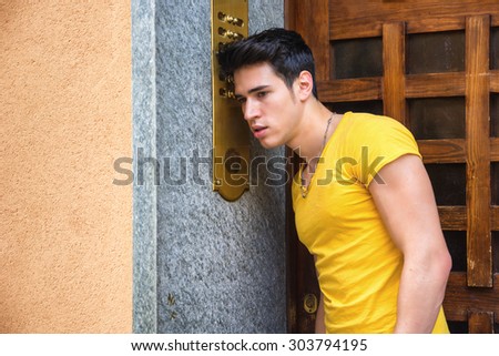 Handsome stylish young man at entrance door on a building ringing doorbell and talking on speaker phone, side view