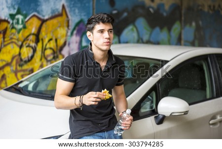 Young handsome man outside his car, standing leaning against it eating food with bottle of water in hand