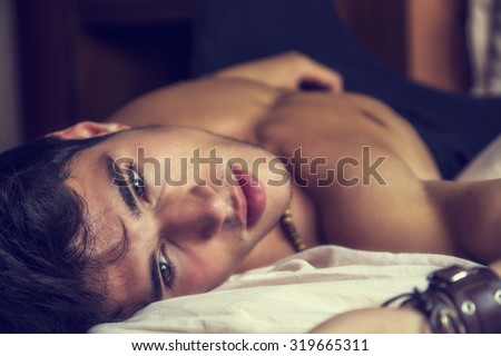 Shirtless sexy male model lying alone on his bed in his bedroom, looking at camera with a seductive attitude