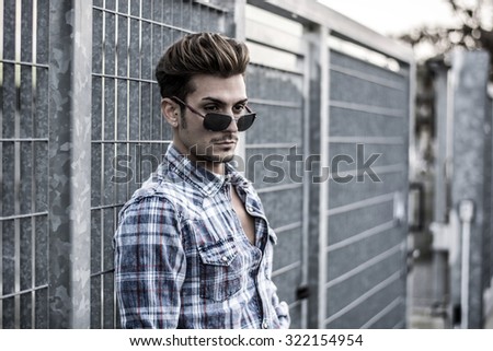Trendy cool young man standing outside, leaning against metal gate, wearing shirt and jeans