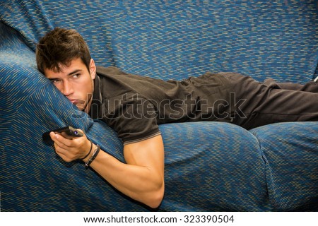 Young bored man sitting on sofa watching television changing the channel with the remote control