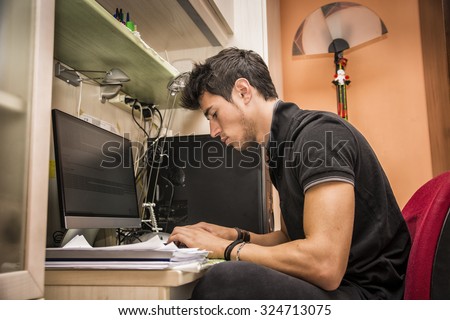 Waist Up Profile of Young Attractive Man with Dark Hair, Sitting at Computer Desk Working on Paper Homework or on His Start-up Business, in Dorm Room
