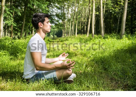 Profile of handsome Young Man During Meditation or Doing an Outdoor Yoga Exercise Sitting Cross Legged on Grassy Ground Alone in Woods