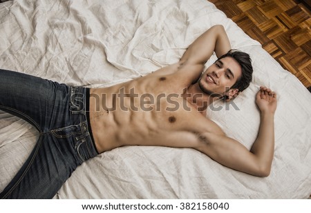 Shirtless sexy young smiling man lying alone on his bed in his bedroom, looking at camera with a seductive attitude