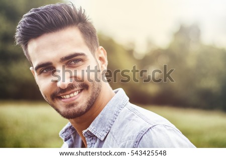 Handsome young man at countryside, in front of field or grassland, wearing shirt, looking away to a side smiling