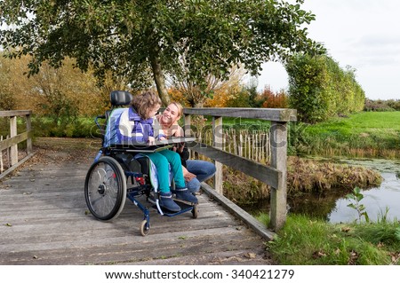 Disabled girl in a wheelchair relaxing by a bridge with a care assistant / Disabled girl relaxing by a bridge