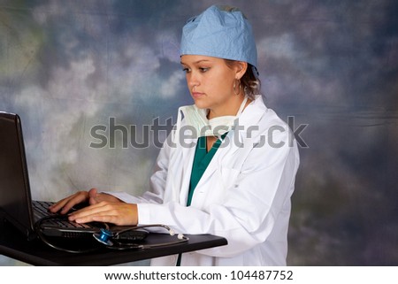 Female medical person in white lab coat, surgery hat and surgery mask around her neck, with a stethoscope around her neck, typing on a laptop computer