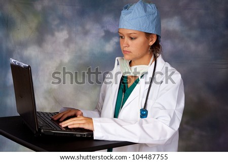Female medical person in white lab coat, surgery hat and surgery mask around her neck, with a stethsocope around her neck, typing on a laptop computer