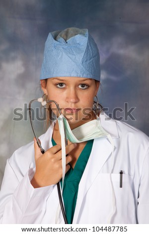 Female medical person in white lab coat, surgery hat and surgery mask around her neck, and holding a stethoscope, looking thoughtfully at the camera with a serious expression