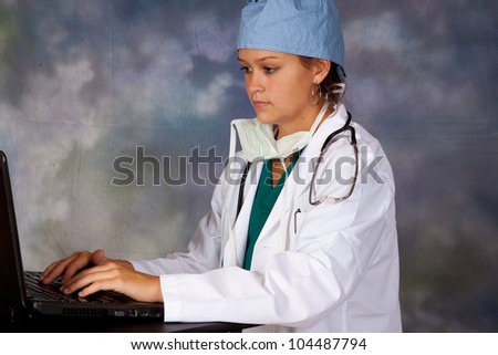 Female medical person in white lab coat, surgery hat and surgery mask around her neck, with a stethoscope around her neck, typing on a laptop computer