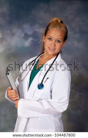 Female medical person in white lab coat, and scrubs, holding a clipboard and looking at the camera with a friendly, happy, smile