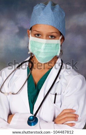 Female medical person in white lab coat, surgery hat and surgery mask around her face, and a stethoscope draped across her shoulders, looking at the camera with her arms crossed