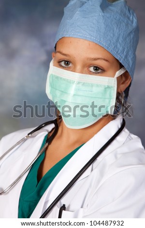 Female medical person in white lab coat, surgery hat and surgery mask around her face, and a stethoscope draped across her shoulders, looking at the camera