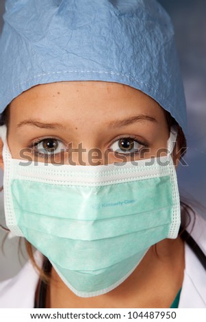 Female medical person in white lab coat, surgery hat and surgery mask around her face, looking at the camera